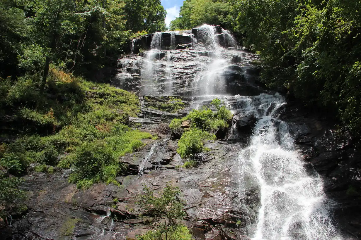 Waterfall cascading over tiers of dark rock surrounded by green summer forest