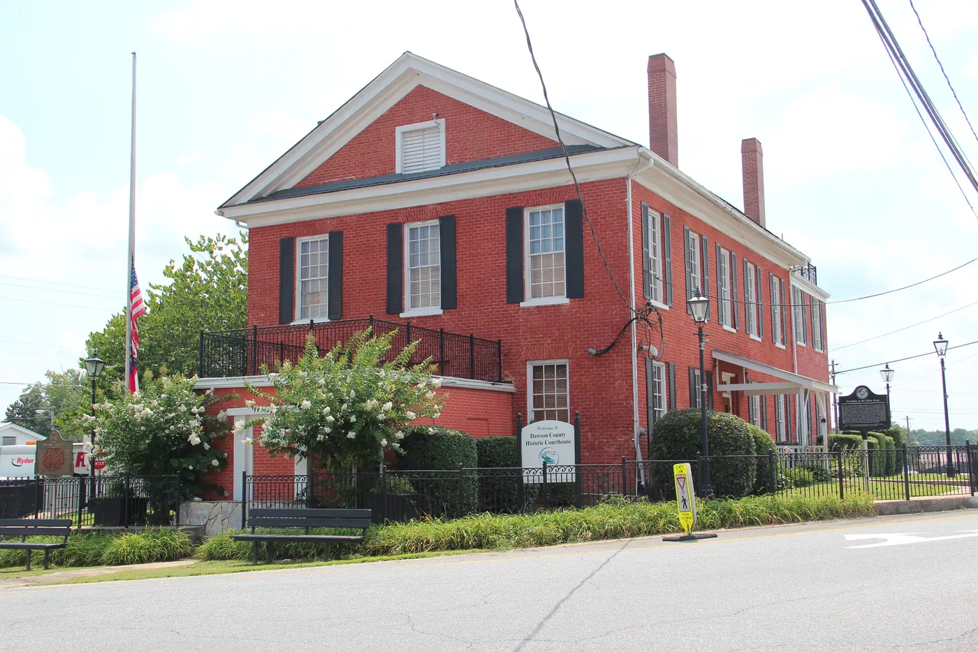 Two-story red brick historic courthouse with black shutters, an iron fence, flowering crape myrtles and an American flag