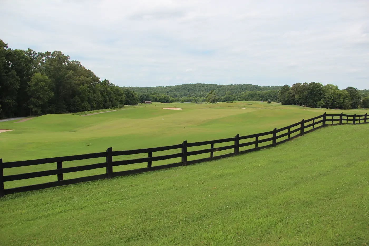 Black wooden fence curving beside a broad green golf fairway with a bunker, tree line and low hills under an overcast sky