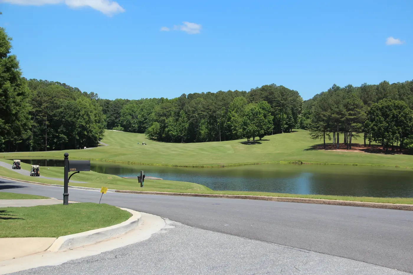 Golf fairway rising behind a pond lined with pine trees, seen from a curving neighborhood street with a mailbox and golf carts on the cart path