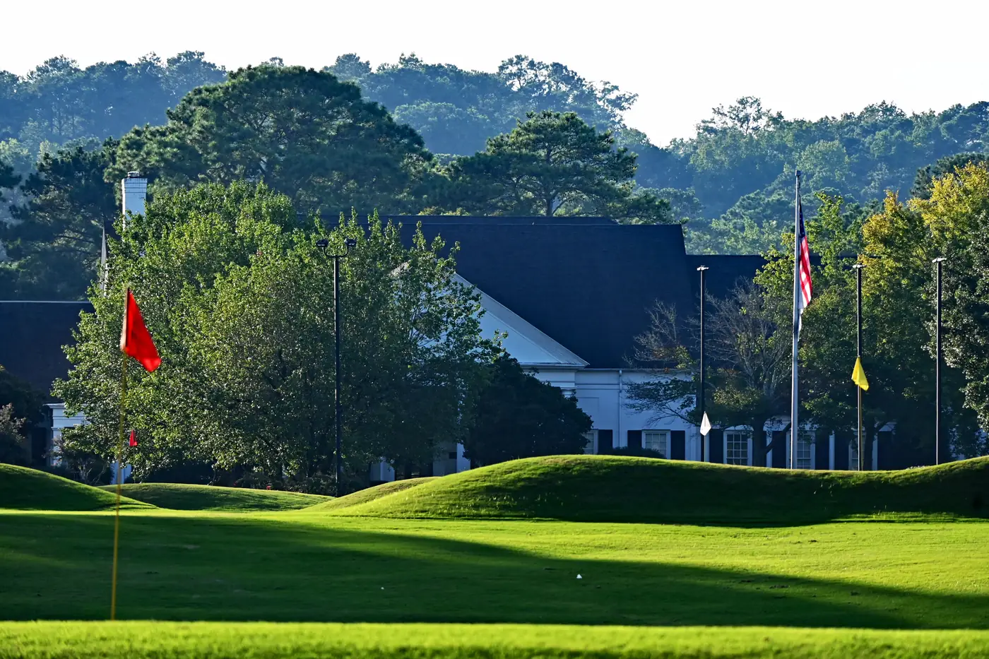 Sunlit putting green with a red flag in front of a white clubhouse, flagpoles and tall trees