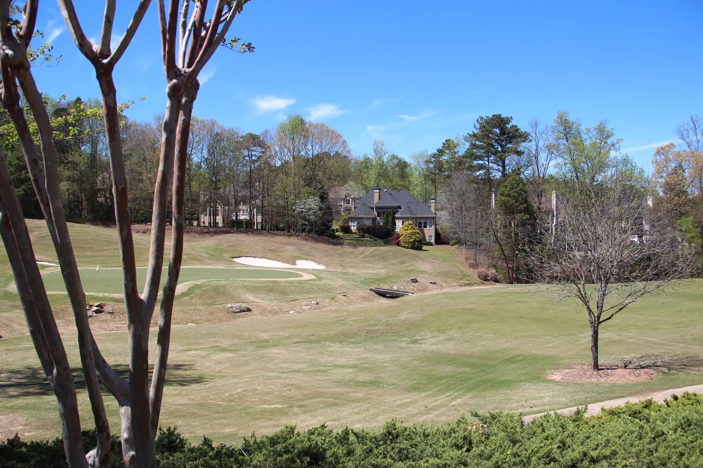 Early-spring golf hole with dormant grass, a sand bunker and a footbridge, with a large house among trees beyond