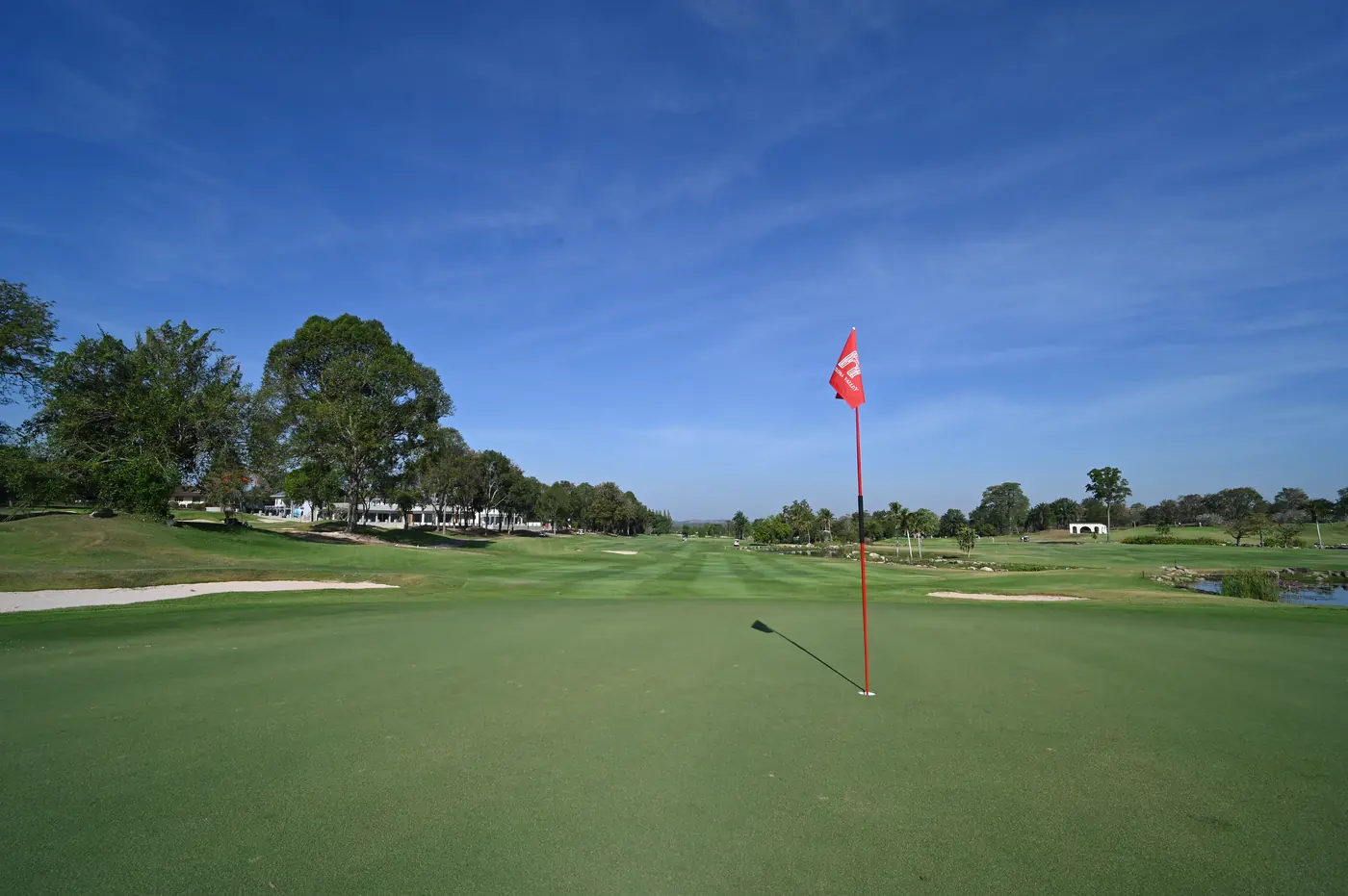 Low view across a putting green to a red pin flag casting a shadow, with fairways, bunkers and trees under a deep blue sky