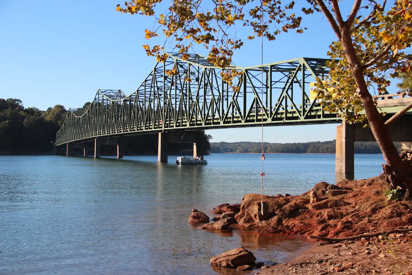 Green steel truss bridge spanning Lake Lanier, with a pontoon boat beneath it and red clay rocks and an autumn tree on the shore