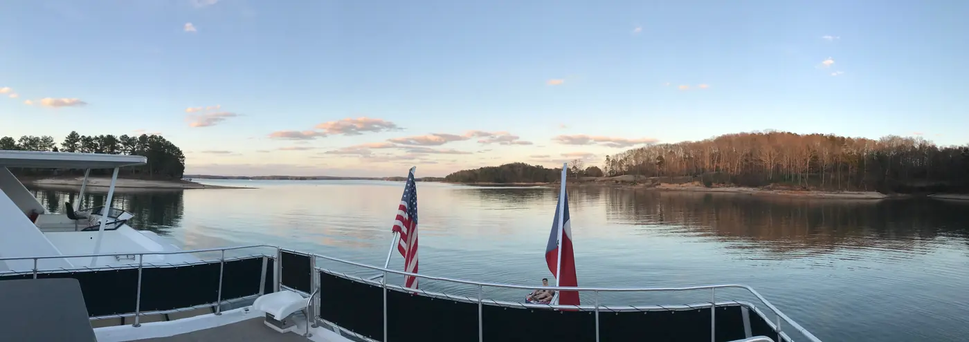Panoramic view from the deck of a houseboat on Lake Lanier at dusk, with American and Texas flags on the railing and a wooded shoreline across calm water
