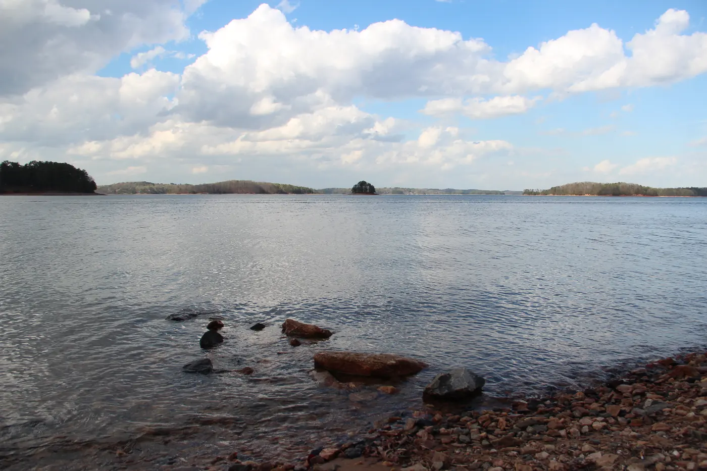 Rocky Lake Lanier shoreline in the foreground with open water, small wooded islands and white clouds beyond